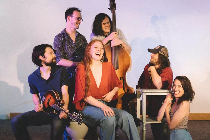 Six people, one standing with a cello, sitting and laughing