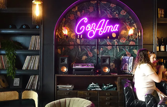 The words "Con Alma" are lit up in a neon sign hanging on the wall behind two women sitting on bar stools in front of the bar