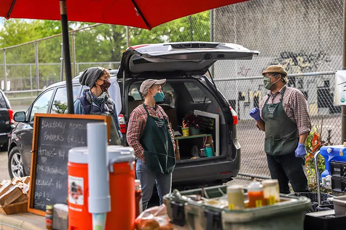 Three adults wearing masks and standing in front of a vehicle with the trunk open as they unload supplies onto their farmers market stand