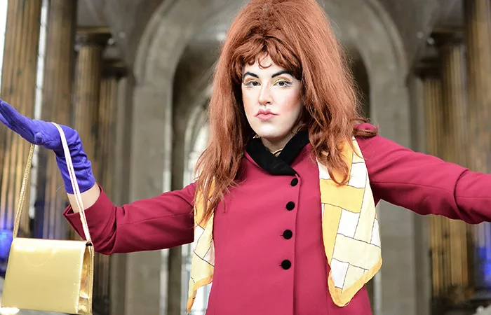 A woman wearing a large red wig poses for the camera in a magenta jacket with black trim, a yellow patterned scarf, purple gloves, and a yellow satin bag.