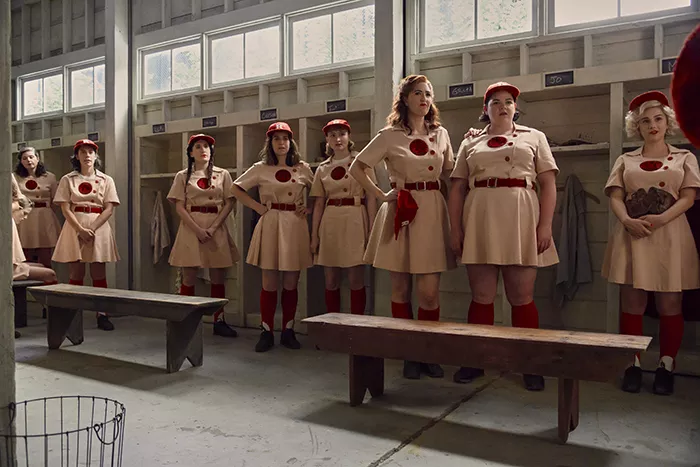 A group of women in tan and red baseball uniforms stand in a locker room, all looking to their left.