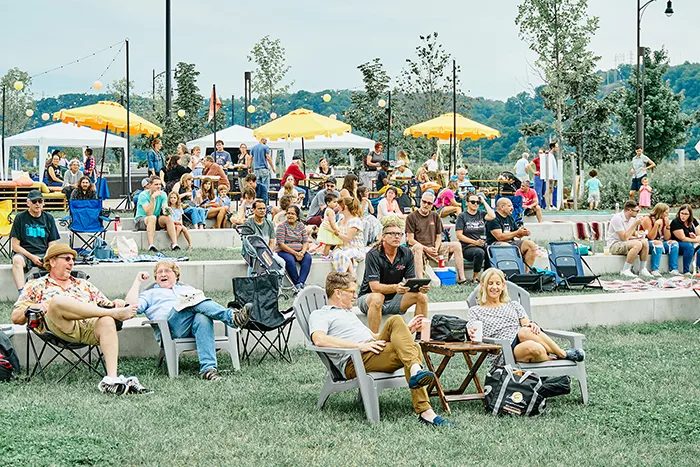 A large crowd of people sit on an assortment of lawn chairs on a grassy area, surrounded by trees, stringed lights, and beach umbrellas