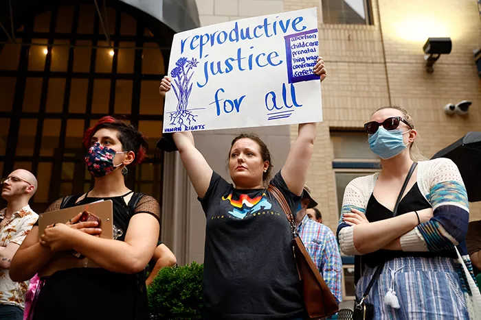 A woman holds up a sign that reads, 
"reproductive justice for all" in between two other people wearing masks with their arms crossed