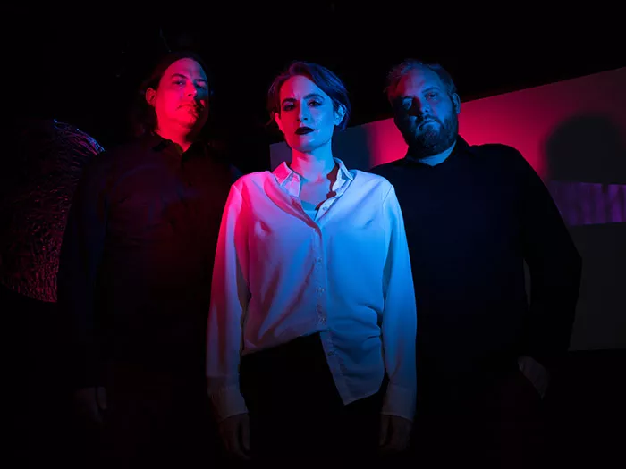 Three people stand looking at the camera with eerie dark and pink lighting