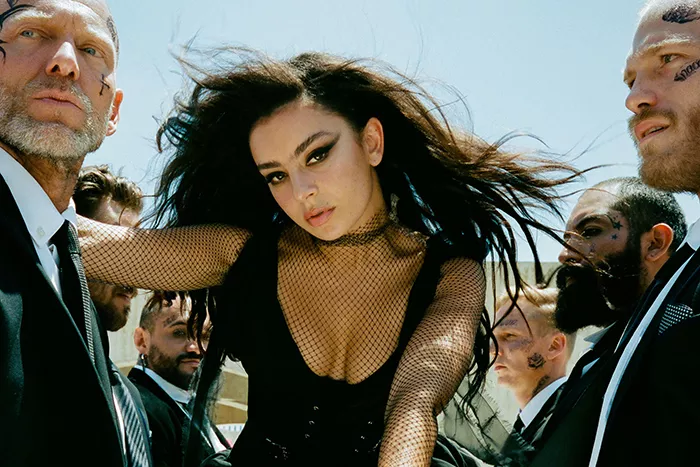 Music artist Charli XCX wears a low-cut, black mesh bodysuit and is surrounded by burly, tattooed men.