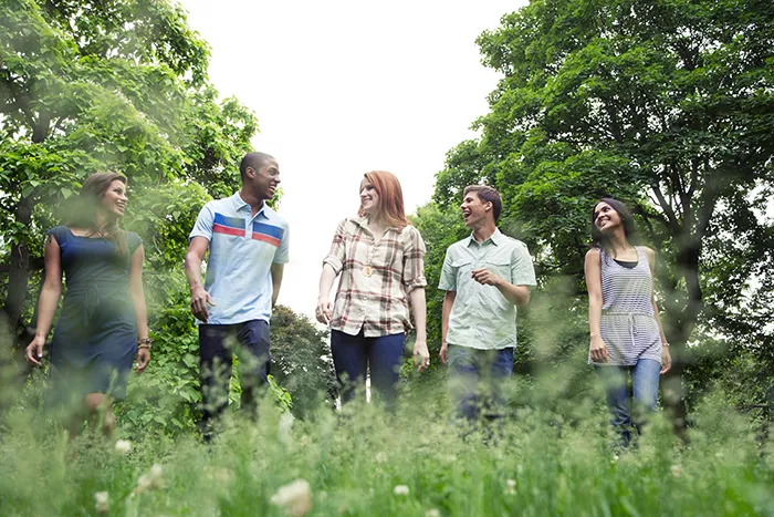A row of smiling teenagers walk through the outdoors with brightly colored green grass in front of them and trees behind them