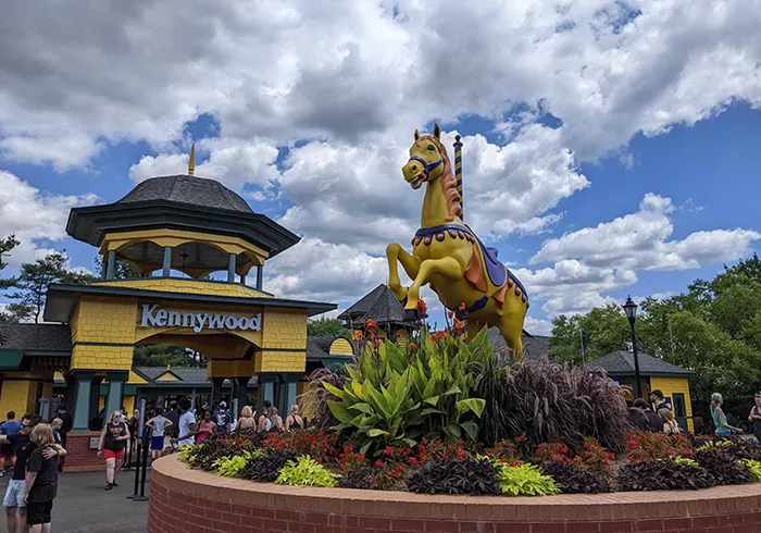 A crowd of people walk through a yellow arched entryway marked "Kennywood" as a yellow carousel horse, surrounded by plants, is displayed in the foreground