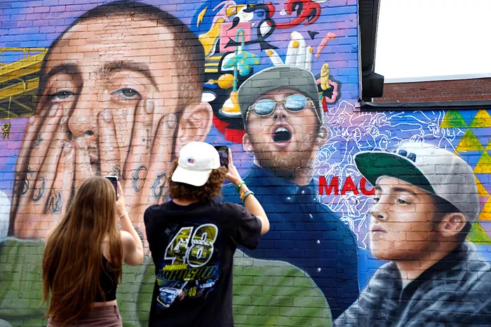 Two people hold up cell phones as they take close-up photos of a large mural that's painted with multiple illustrations of late Pittsburgh musician Mac Miller