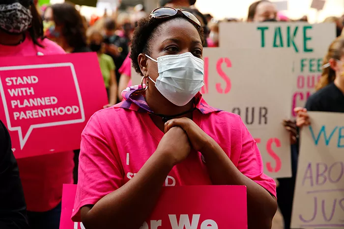 A woman wears a mask and a Planned Parenthood T-shirt in front of a crowd of people holding up signs for abortion rights