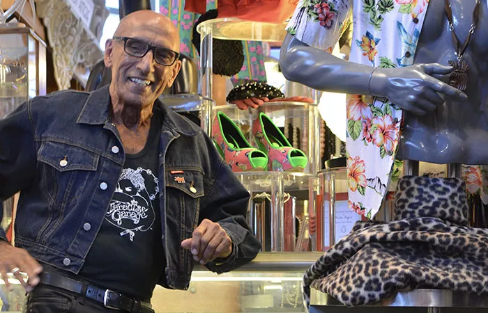 A smiling man in black jeans, black belt, black-and-white T-shirt, and a dark denim jacket leans against a display case that shows off bright neon green and pink shoes and other accessories