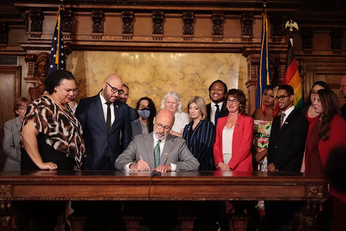 A man sits at desk and signs paper while a large group of people surround him smiling