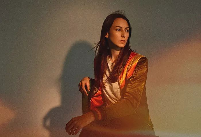 Woman with long brown hair, wearing a brown jacket with orange and yellow stripes, sits on a chair and looks off to the left of the camera