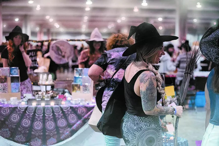 A crowd of people wearing witches' hats, browsing through boots selling various wares including candles and broomsticks