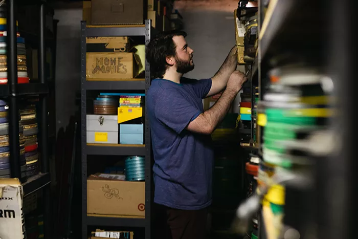 A dark-haired bearded man looks through film reels and boxes of film supplies on shelves in a basement