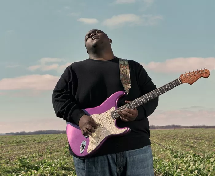 Man in jeans and a black long-sleeved shirt stands in a field, holding a purple guitar and looking upwards in front of a blue sky