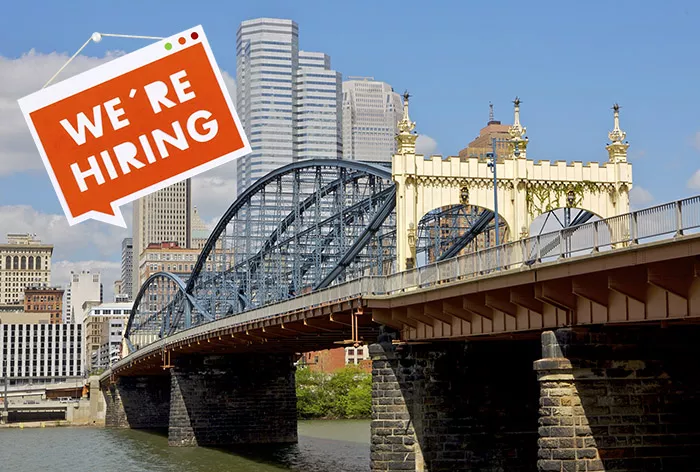 A bridge goes over a river in front of the Downtown Pittsburgh skyline. A sign that says "we're hiring" is Photoshopped on top of the photo