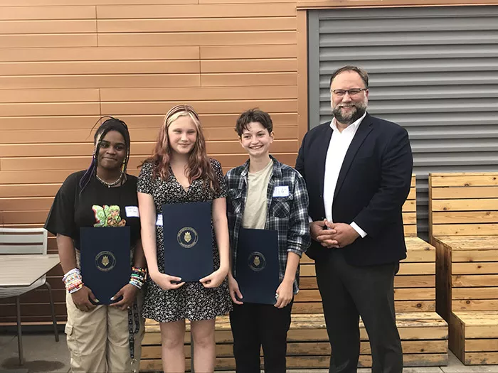 Three smiling teenagers stand next to a smiling man. Each kid is holding up a folder with a city of Pittsburgh seal on the front