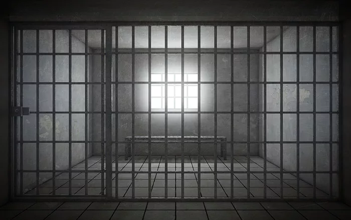 Empty prison cell showing bars, an empty bench, and light coming in through the window