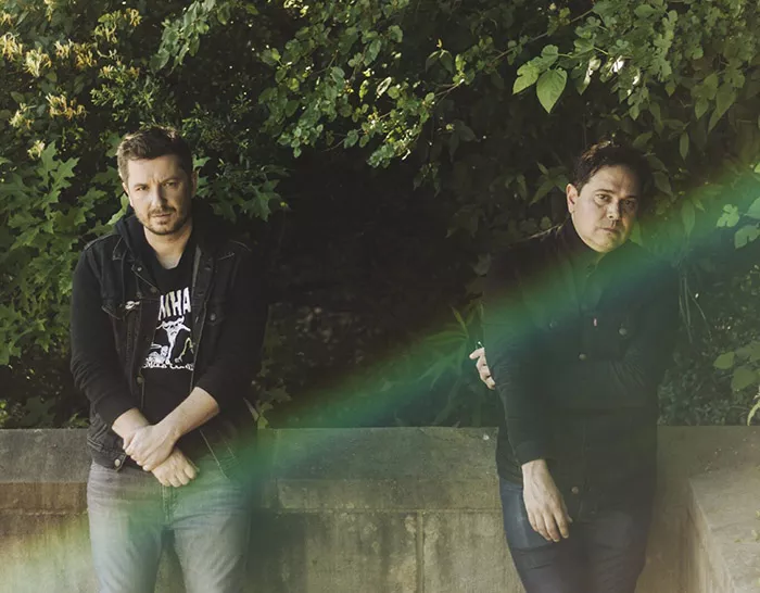 Two people in black shirts and jeans stand in front of a wall and green trees. Both are looking at the camera with solemn expressions