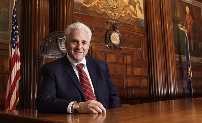 Smiling gray-haired man in a blue suit, white shirt, and red and gold striped tie sits at a wooden desk beside an American flag