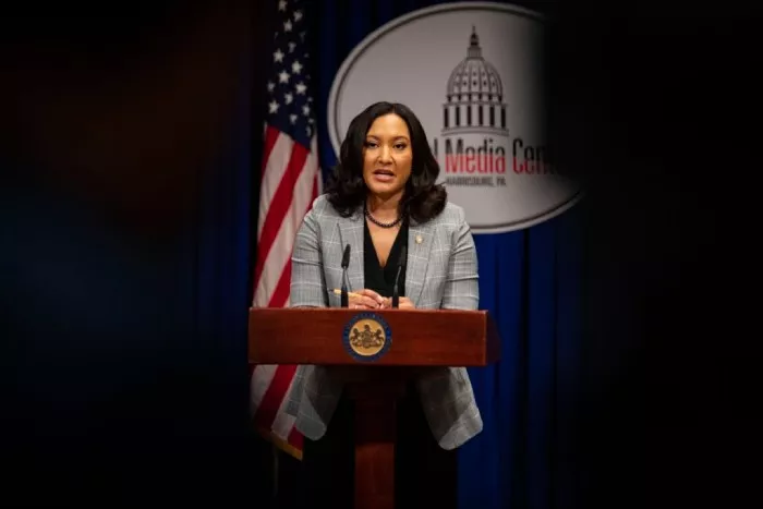 Woman standing and speaking at a podium in front of a blue curtain and an American flag