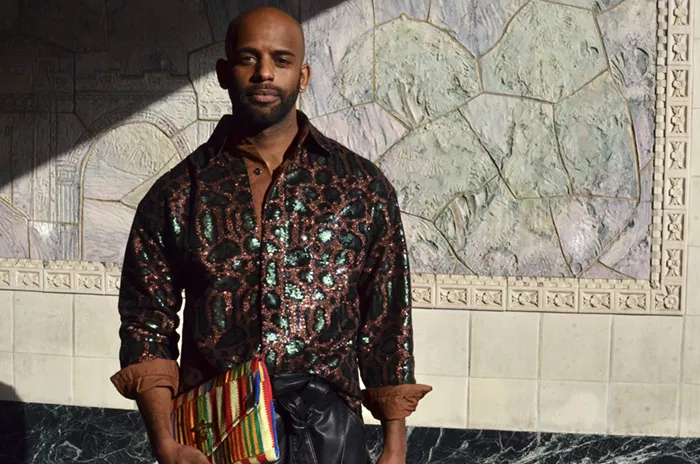 A man, in a shiny brown and blue patterned long-sleeved shirt, looks at the camera, confidently holding a multi-colored purse