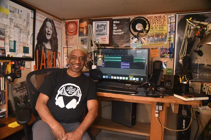 A Black man sits at a desk in front of a radio studio set up.