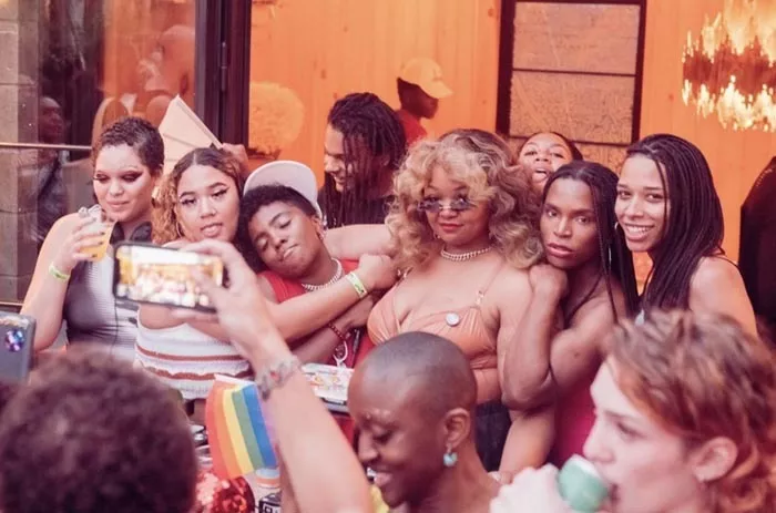 A group of Black DJs and performers wearing club outfits pose during an outdoor event.