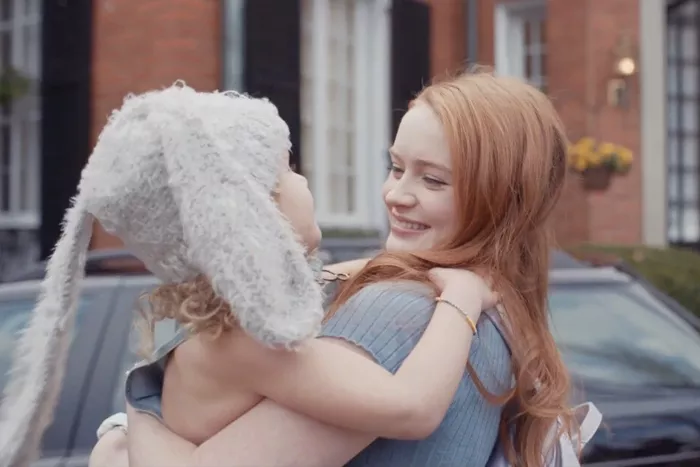 A girl with red hair smiles as she holds a child wearing a hat with bunny ears.