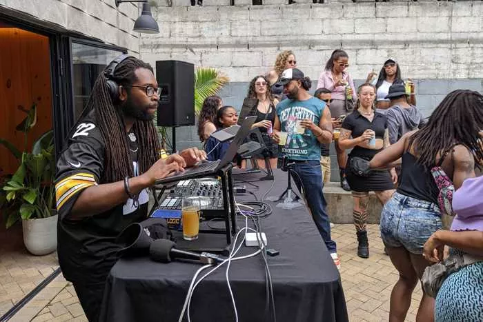 A Black DJ leans over his setup as people dance around him.