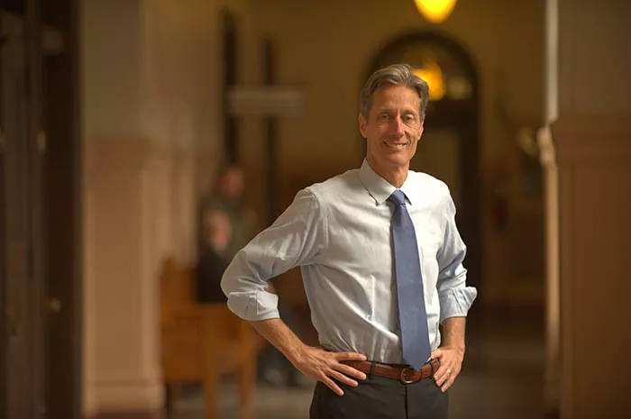 A tall white man wearing a button-down shirt and tie stands in a lobby with his hands on his hips.