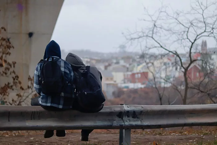 Two people sit together on a guardrail. One person leans their head on the other's shoulder