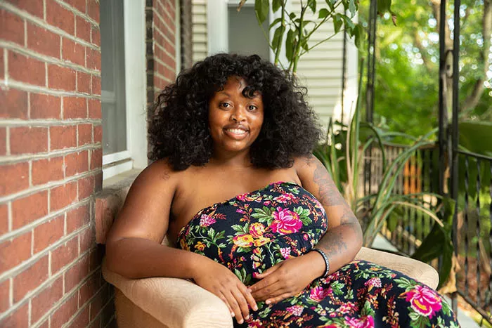 A young Black woman in a bright floral dress smiles at the camera as she sits on a porch.