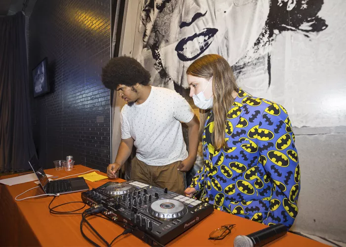A Black teen with an afro and wearing a white T-shirt, and a white teen wearing a blazer covered in Batman logos and face mask, stand behind a table with Dj equipment.