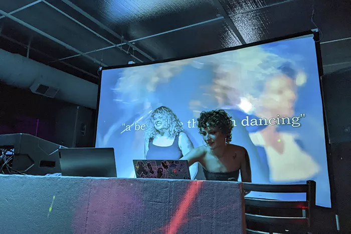 Two women dance near a DJ deck as a projection plays behind them.