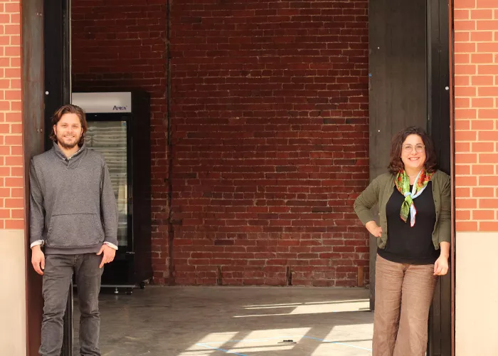 A man and woman stand across from each other in front of a brick facade as they pose and smile for the camera.