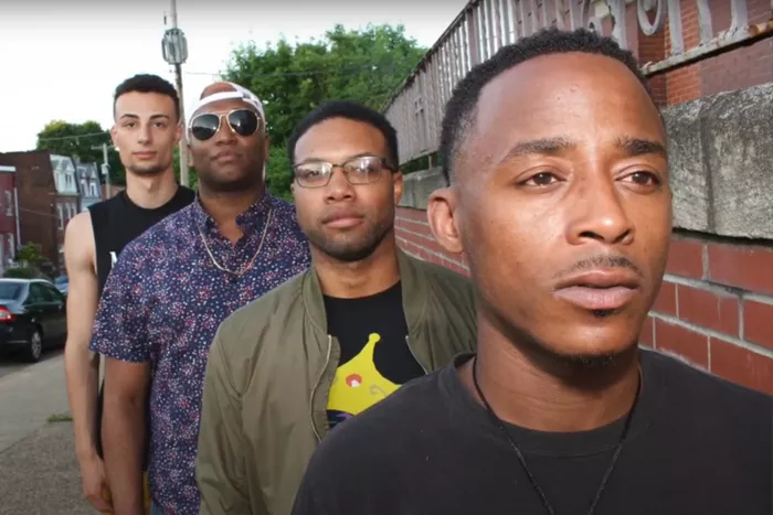 Four Black men pose in a row on along the side of a building, each one with a different look.