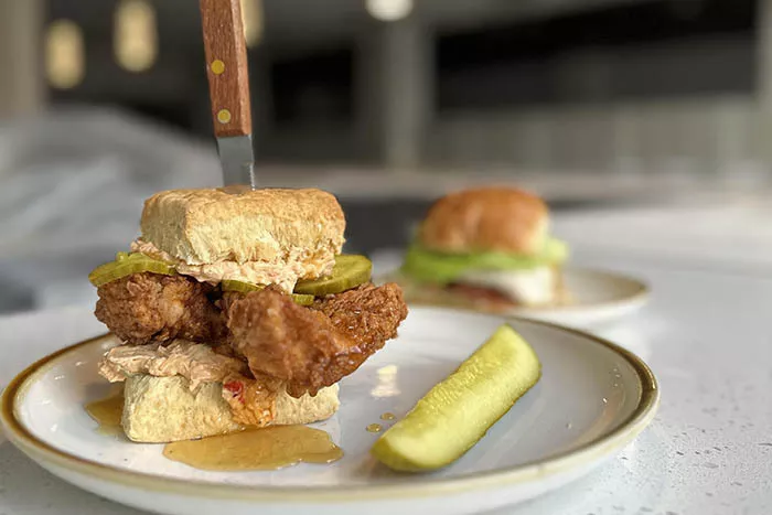 A knife is stuck through a biscuit sandwich with a large piece of fried chicken, pickles, and honey drizzle. It sits on a plate next to a pickle.