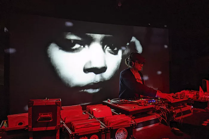 A DJ awash in red light plays as a the projected image of a face shines behind him.