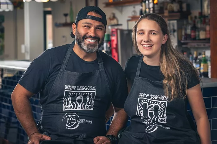 A man wearing a backward black baseball cap and a woman with long, wavy blonde hair pose together wearing black T-shirts that read "Best Buddies."