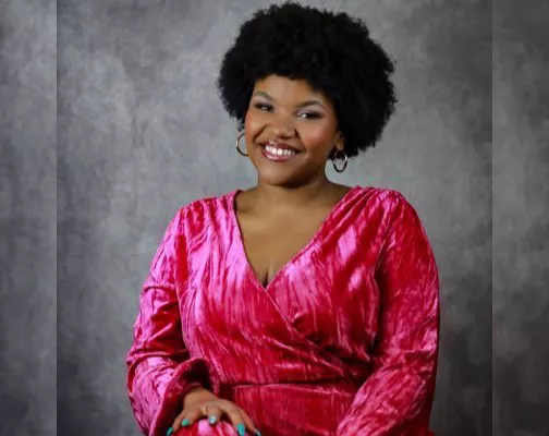 Ashley Comas has puffy natural hair and wears hoop earrings and a pink satin dress.