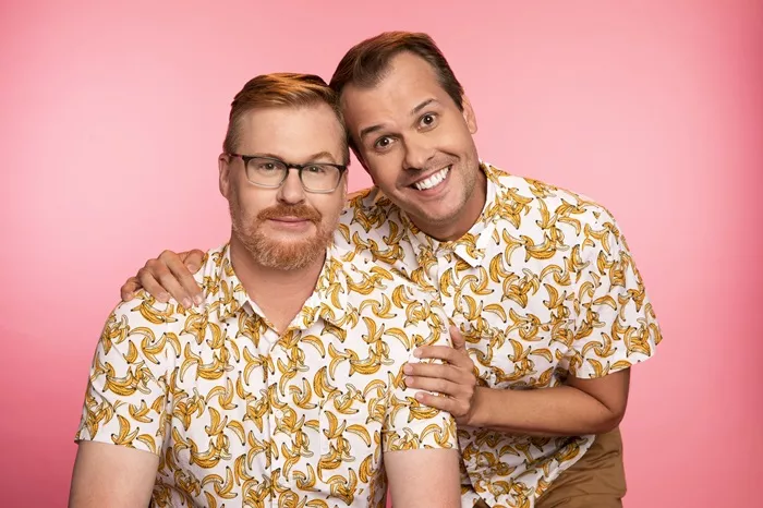 Two men wearing white button-down shirts featuring a banana print pose smiling in front of a pink background.