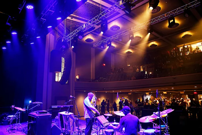 Blue lights shine down on a band playing on stage, with audience members standing in front of the stage