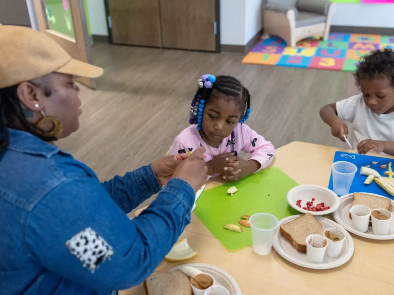 Two young kids with braided hair and dark skin listen as a woman peels a banana at a table with sandwiches on plates