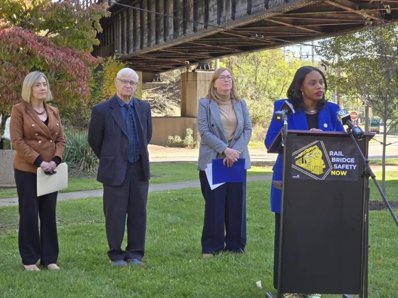 A woman in a blue blazer and formal pants stands beneath a rusted rail bridge with other officials at a podium