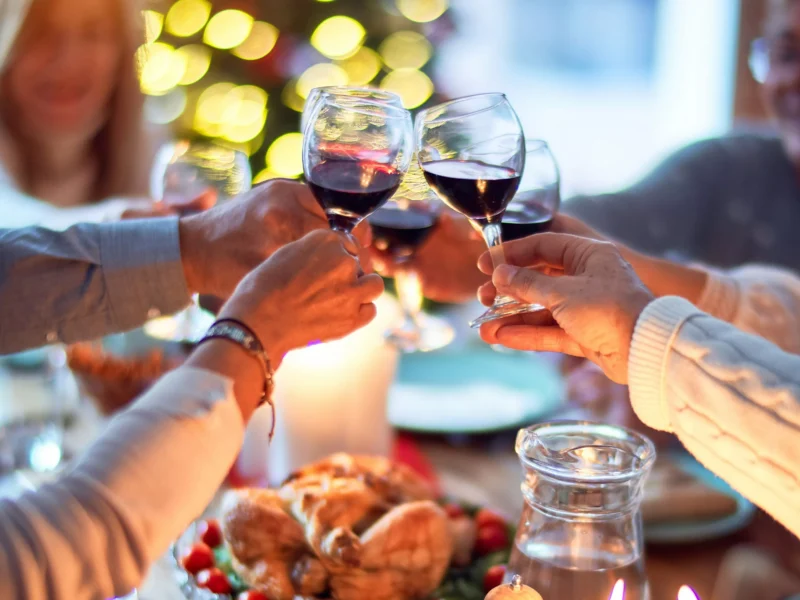 Hands are extended over the Thanksgiving table to clink wine glasses. Out-of-focus faces smile in the background