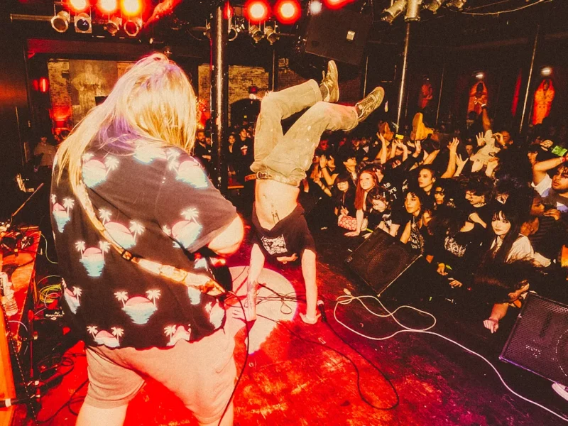 Tattooed members of a punk band play a crowded room as the vocalist does a handstand