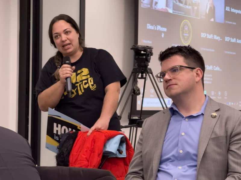 A woman holding a sign and sweatshirt speaks near a clean-shaven man in a sport coat