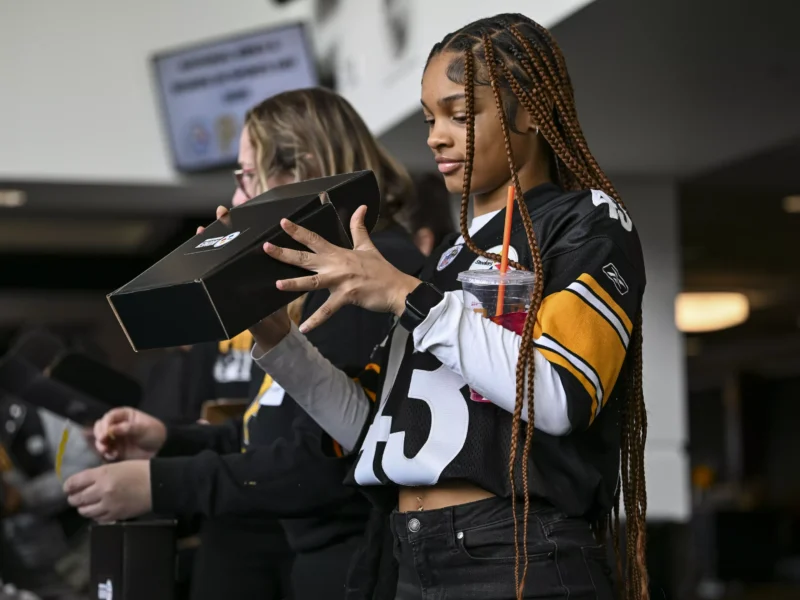 A woman with braided hair wearing a Steelers jersey inspects a box containing a sports bra and swag from local teams