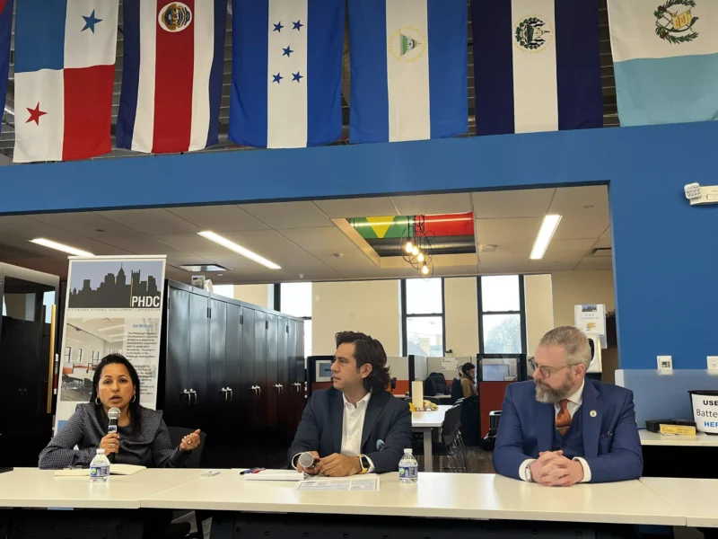 Three speakers sit in formal clothing beneath a display of Central American flags